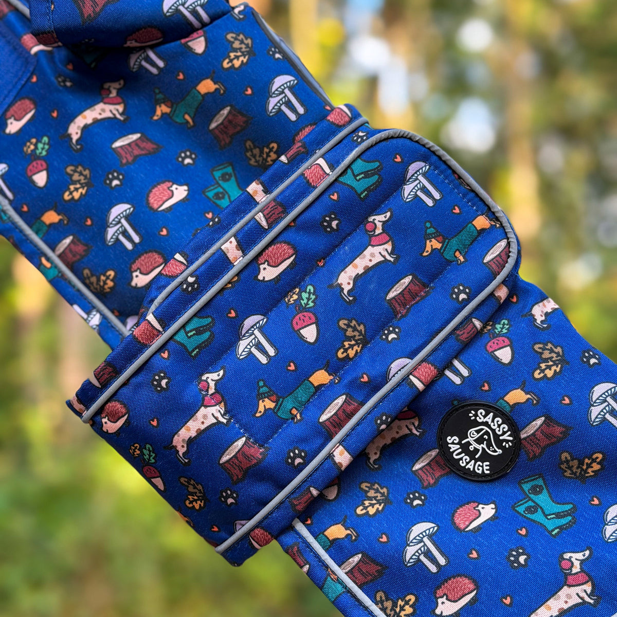 A close-up of a blue fabric dog leash with a whimsical dog and mushroom print, featuring white piping and a black circular 'Coastal Garage' logo patch. The background is a soft, blurred outdoor forest setting.